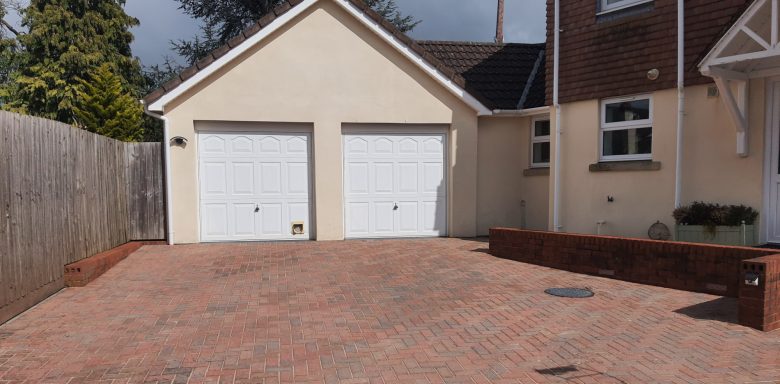 A driveway with red brick paving leading to a double garage with white doors.