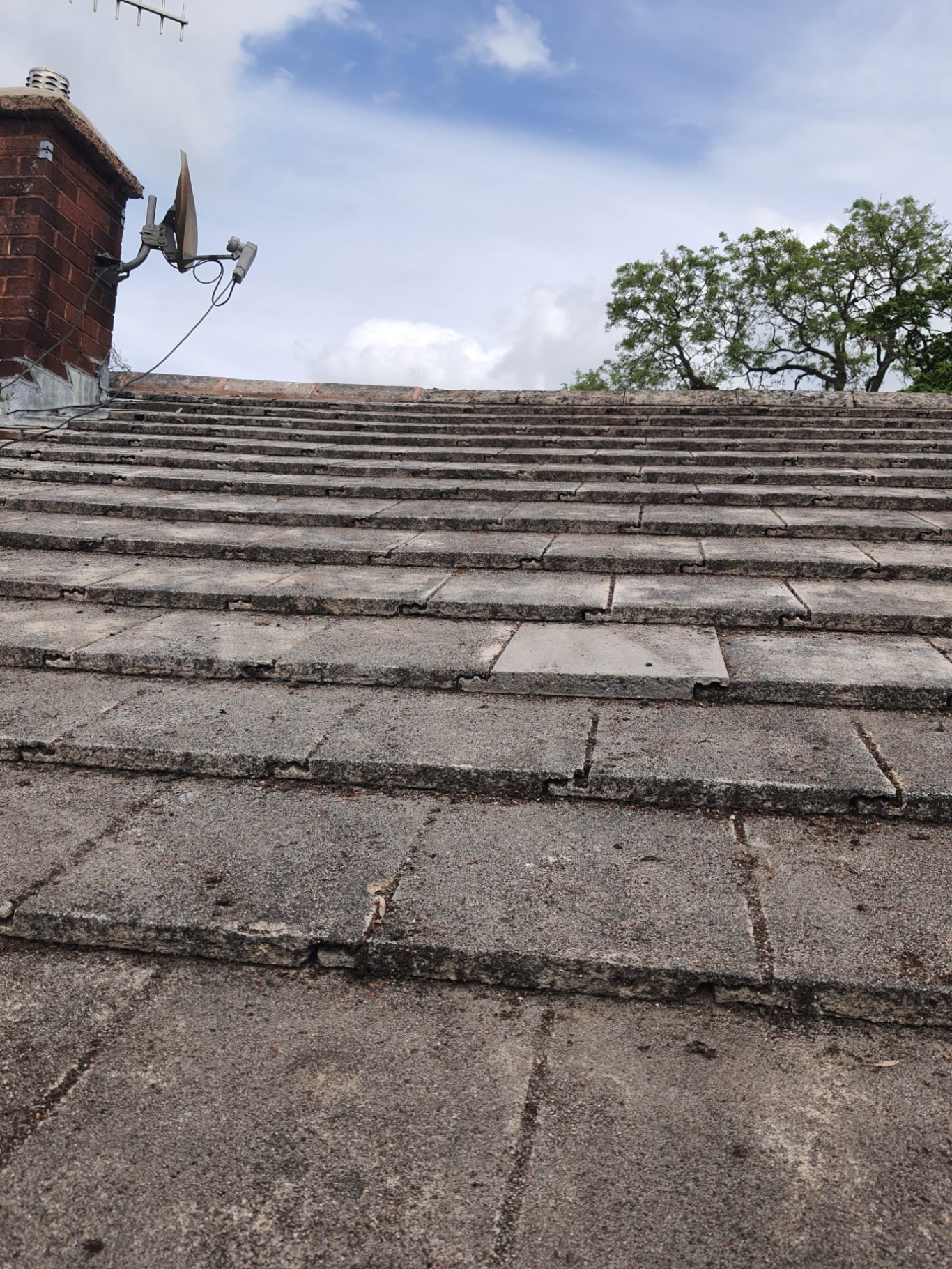 Moss Removal Weathered, sloped roofing tiles leading upwards, with a cloudy sky above and trees in the distance.