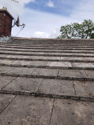 Weathered, sloped roofing tiles leading upwards, with a cloudy sky above and trees in the distance.
