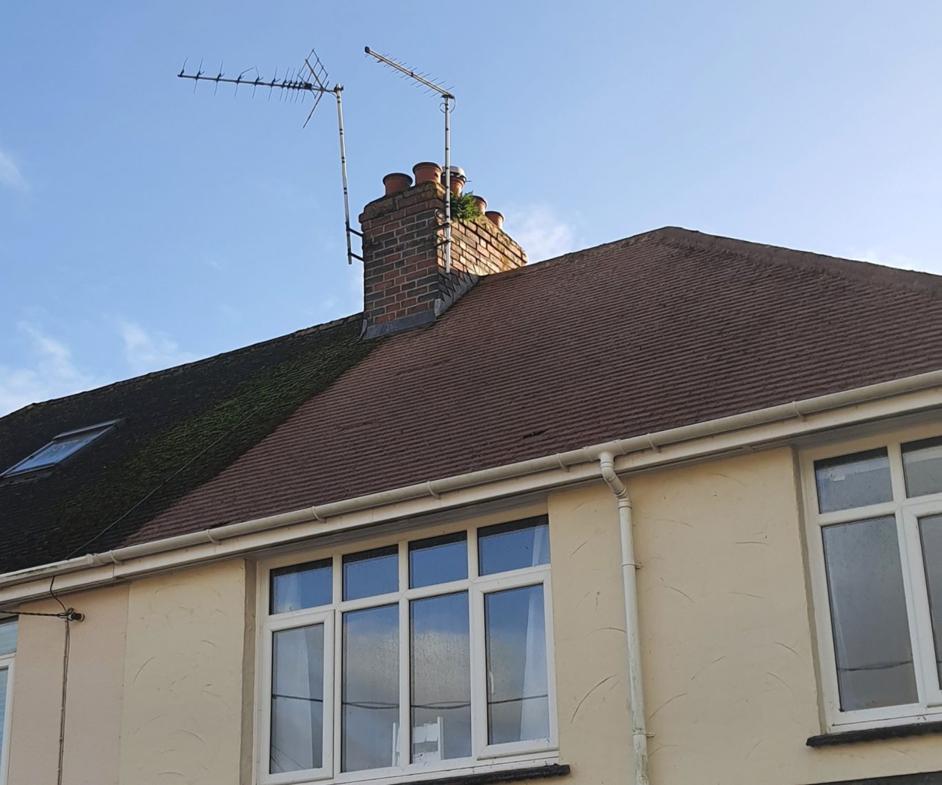 Moss Removal View of a house roof with a chimney and TV aerials against a blue sky.