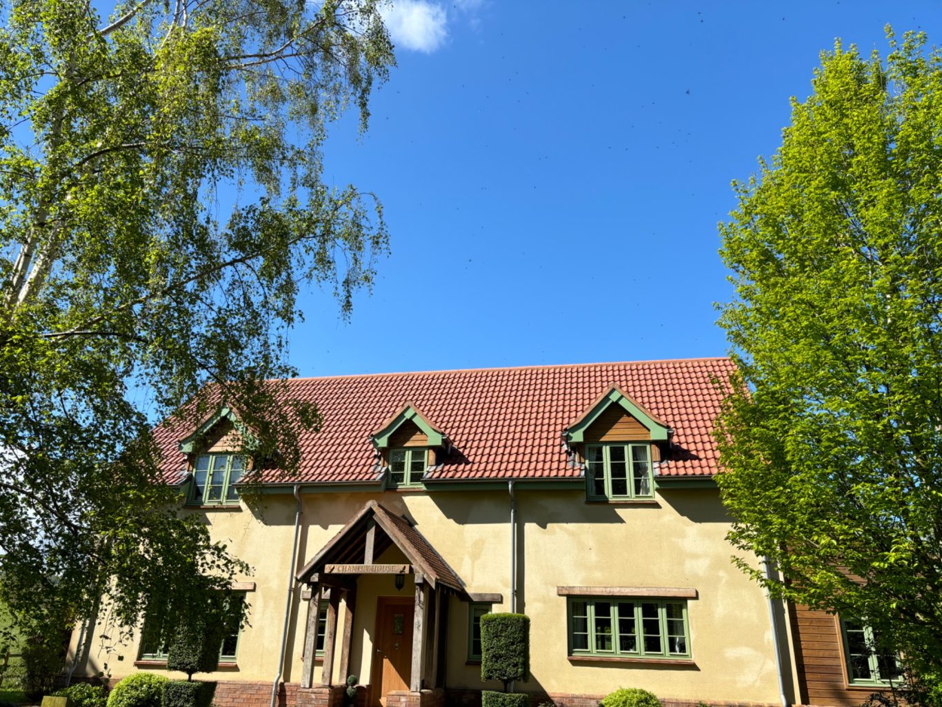 Roof Cleaning In Somerset Two-storey house with green window frames, surrounded by trees under a blue sky.