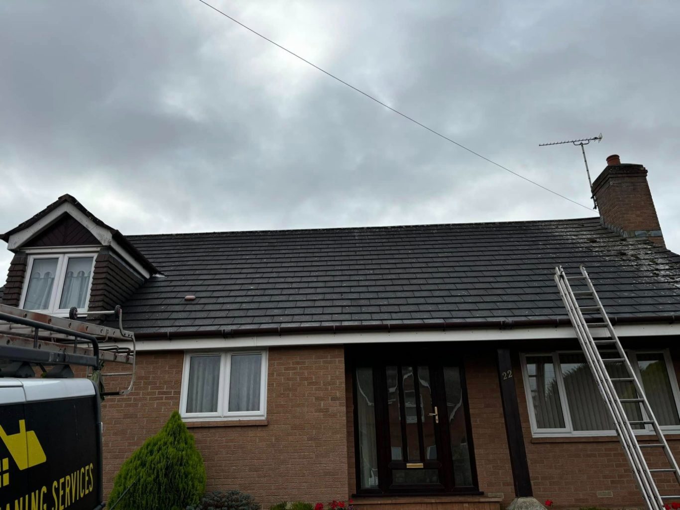 Roof Cleaning A house roof with grey tiles under a cloudy sky, with a ladder leaning against it.