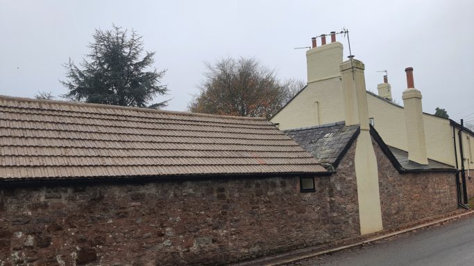 Stone wall with a tiled roof, lead to a yellow house with multiple chimneys.
